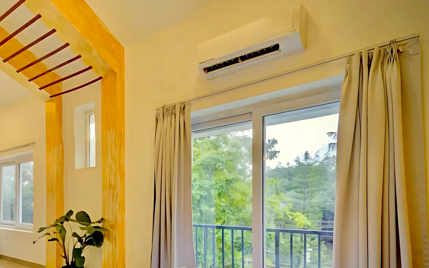 Ar-condicionado split branco no alto da parede de um quarto claro, sobre a porta de vidro da varanda, com árvores lá fora